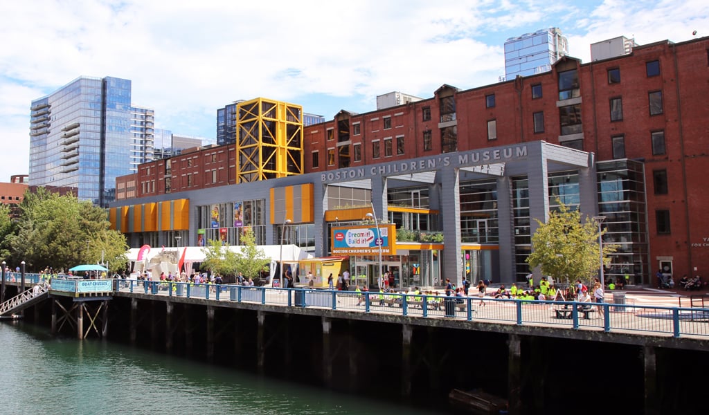 The Boston Children's Museum featuring a distinctive brick and glass exterior with a large yellow structure, set against a backdrop of modern skyscrapers.