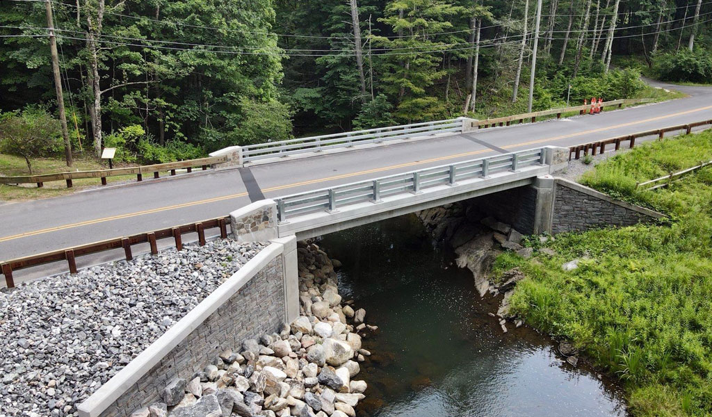Aerial view of a small bridge over a stream, surrounded by green trees and a winding road, in a rural setting.