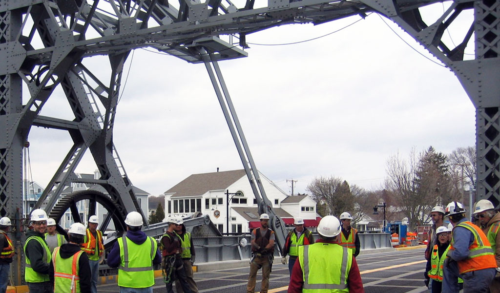 A group of construction workers wearing safety gear under a large metal bridge structure.