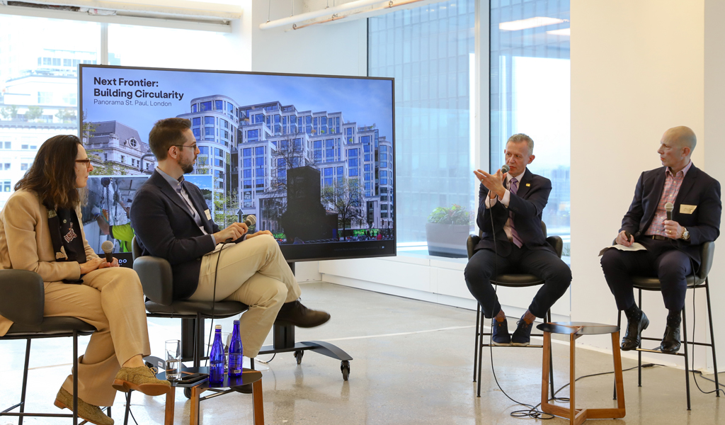 A panel discussion featuring 4 speakers seated in front of a screen displaying building images.