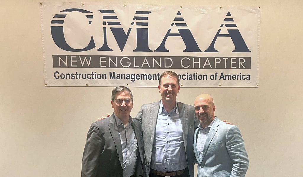Three individuals stand smiling in front of a banner that reads "CMAA New England Chapter, Construction Management Association of America."