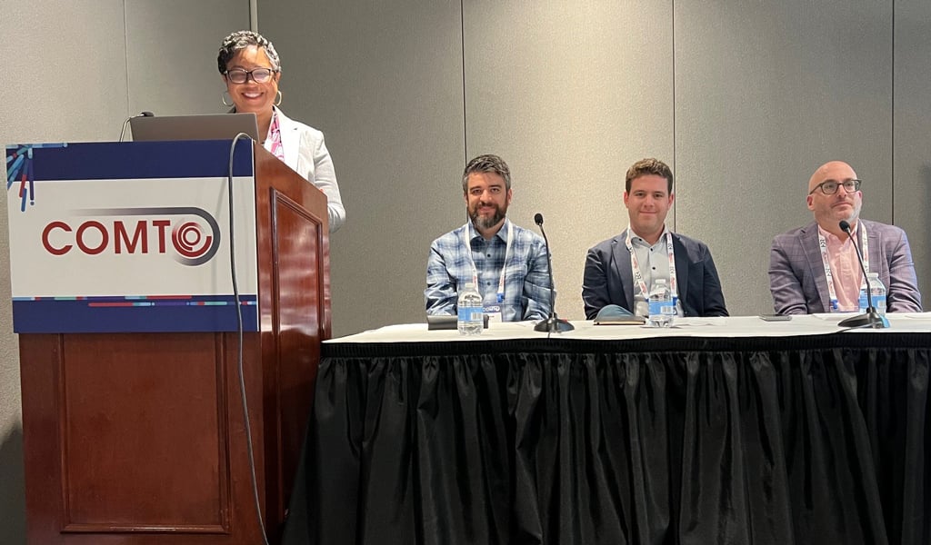 Four VHBers presenting at a conference on a stage, sitting behind a rectangular table with the moderator at the podium.
