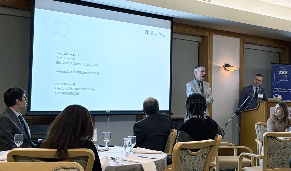 A conference room with attendees seated at tables. Two presenters stand at a podium near a screen. The American Society of Civil Engineers (ASCE) logo is seen on a banner.