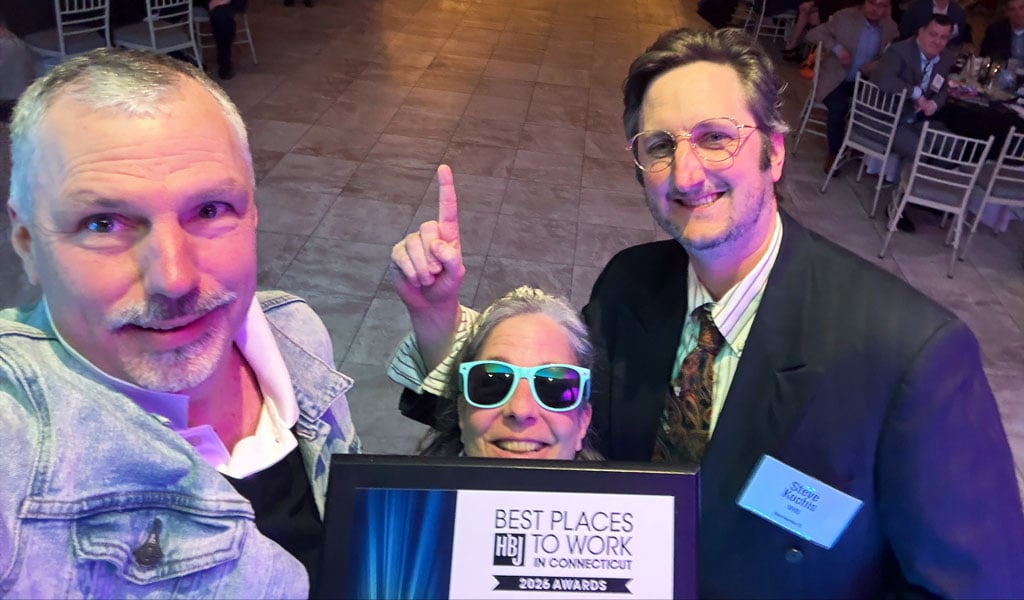 Three individuals on a stage taking a selfie holding a Best Places to Work plaque with a crowd in the background. 