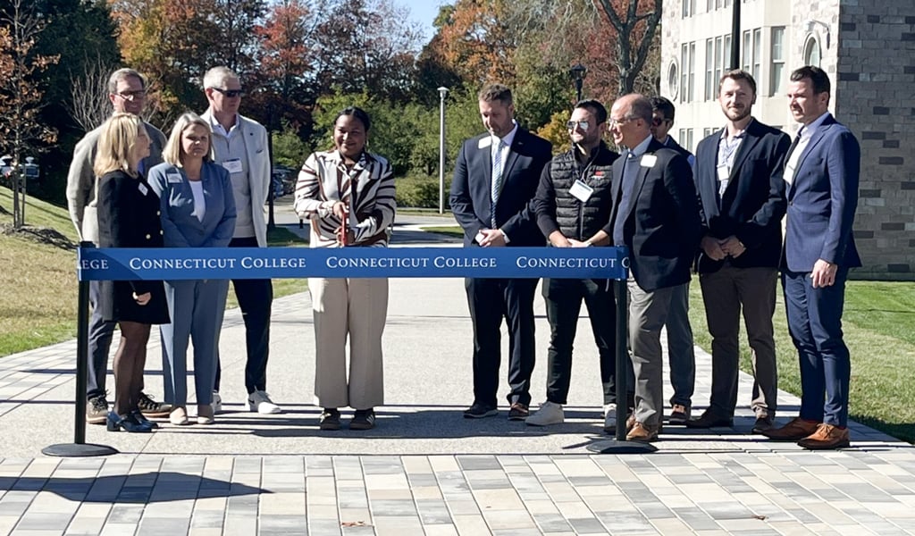 A group of people stands together on a pathway outside a building, with a "Connecticut College" ribbon held in front. One person in the center holds scissors poised to cut the ribbon. 