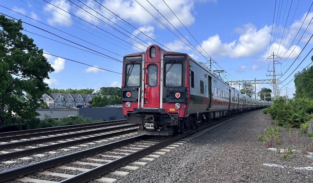 A red and silver Metro-North Railroad train travels along a track on a clear day with scattered clouds. Trees and houses are visible in the background.