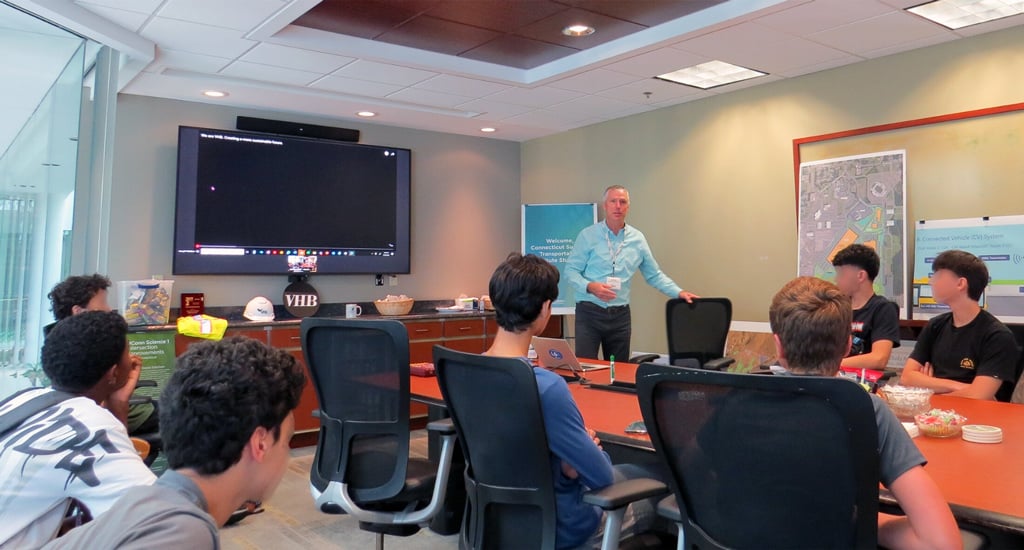 Person standing and speaking to a group of students seated around a conference table in a meeting room with a large screen and map on the wall.