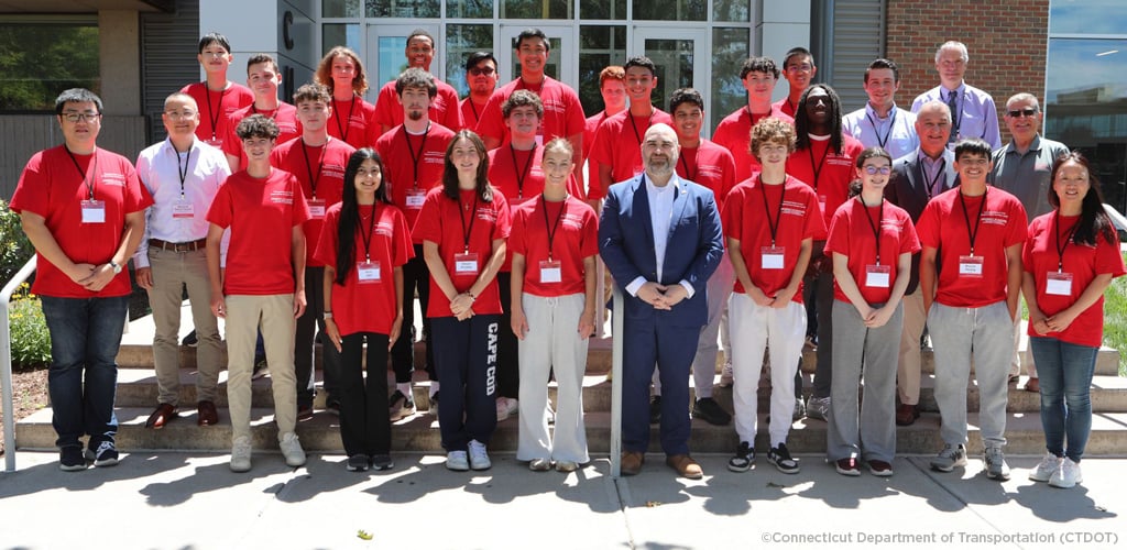 Group of people in red shirts with name tags, standing with a few individuals in business attire, in front of a building with trees and clear sky in the background.