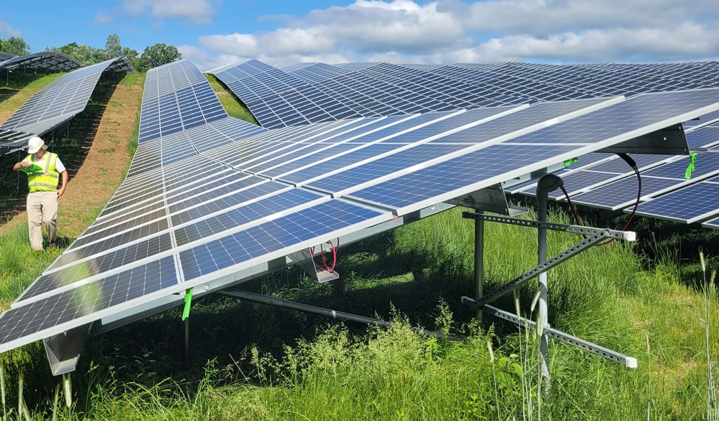 A person in a high-visibility outfit inspects a large solar panel array in a green field under a partly cloudy sky.