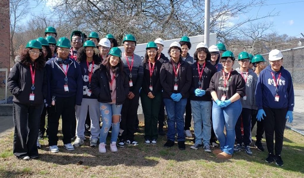 Group of student volunteers wearing green hard hats and blue gloves standing outdoors for a community clean-up event.