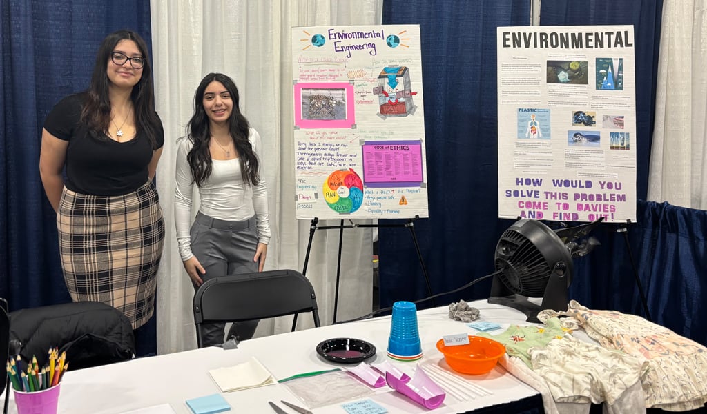 Two students standing in front of a display at the William M. Davies Jr. Career and Technical High School engineering booth, featuring posters and various educational materials on a table.