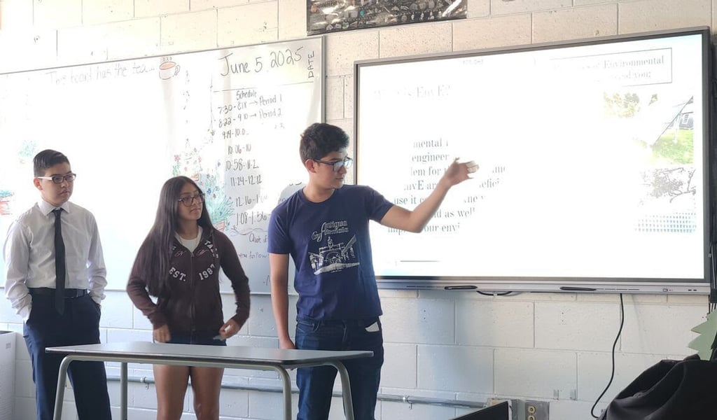 Three students presenting a science project in a classroom, with one student pointing to a data chart on a screen.