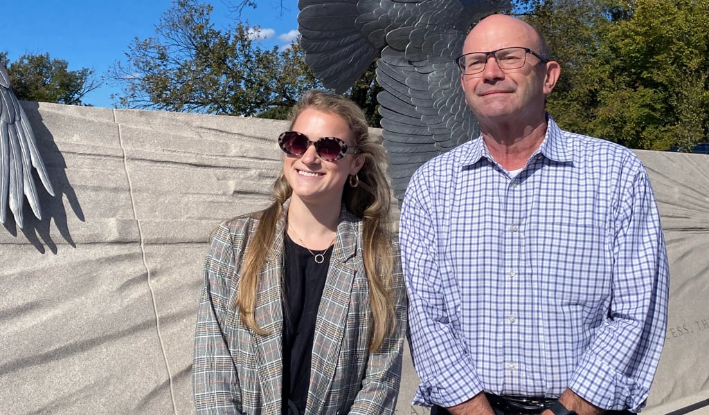 A woman in a plaid blazer is smiling alongside a man in a checked button up shirt in front of a bronze statue of a bird. 