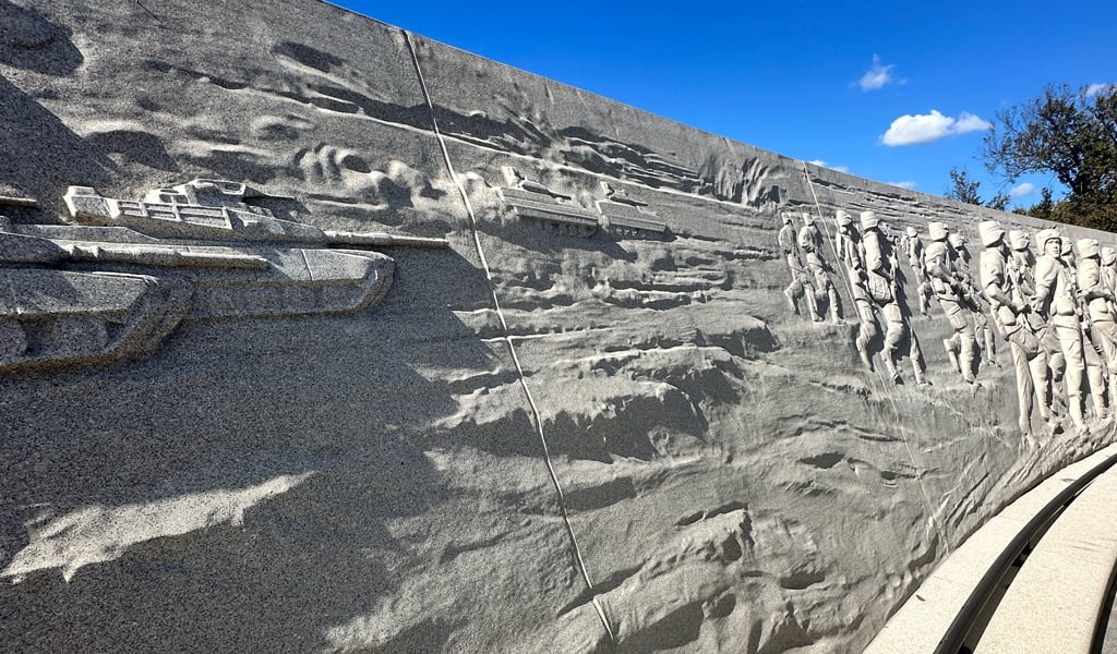 A tan sandstone wall curves around the memorial with sculpted features reliefs depicting the events of the operation.