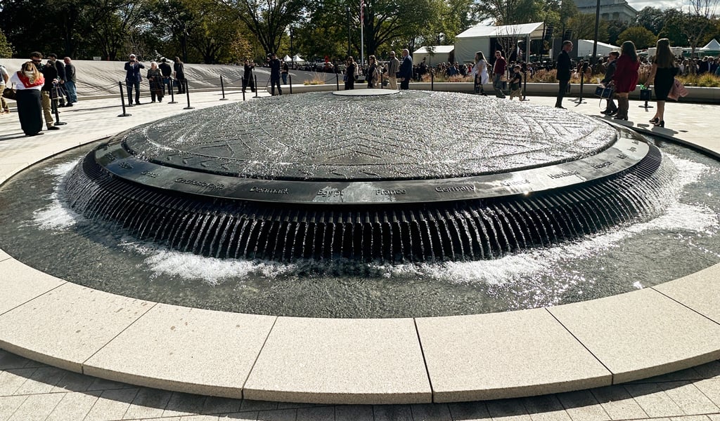 A large bronze shield fountain is at the center of the memorial, surrounded by visitors and blue skies.
