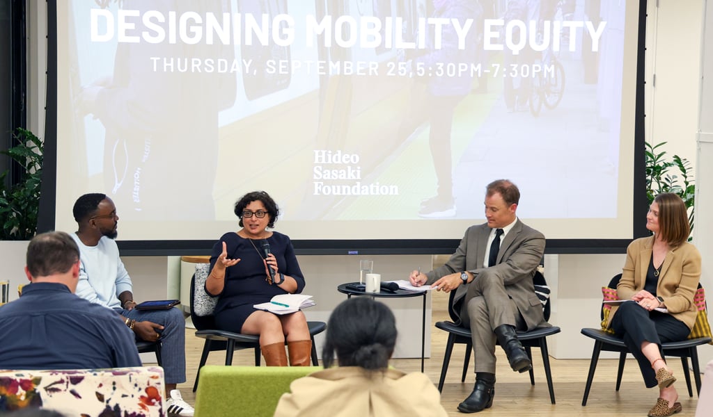 A modern conference room hosts a panel discussion on 'Designing Mobility Equity,' organized by the Hideo Sasaki Foundation. Audience members sit on colorful and contemporary furniture, attentively listening to the speakers.