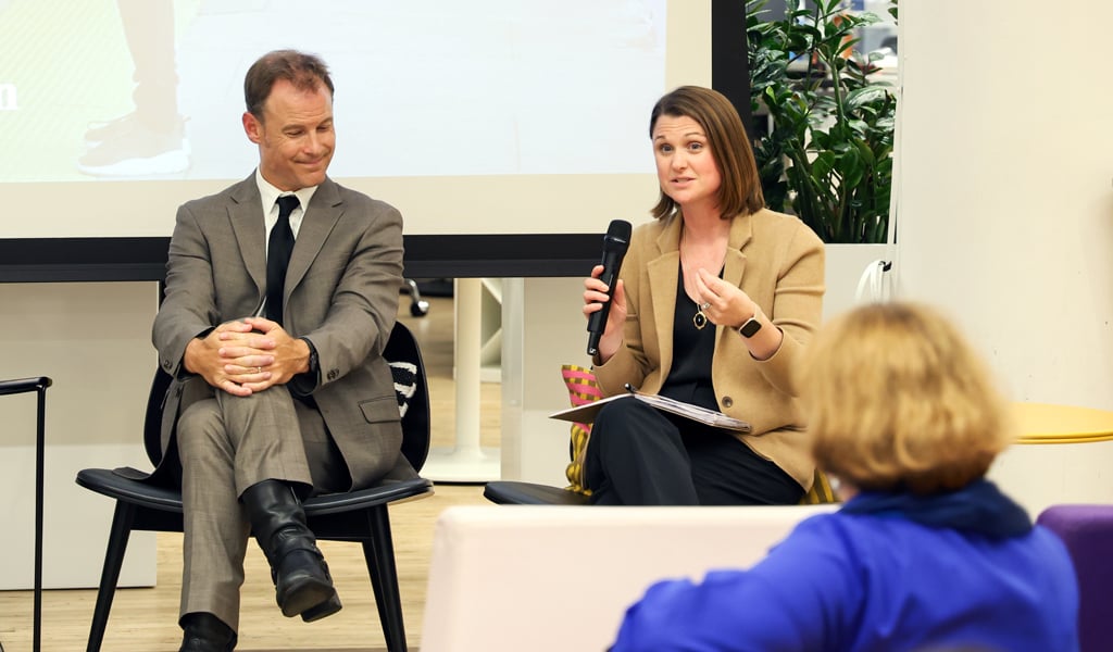 Woman speaking into a microphone during a panel discussion, seated next to a man in a gray suit.