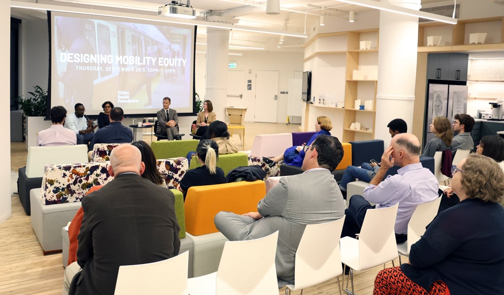 A group of people sitting in a modern conference room, attentively watching a panel discussion. The presentation screen displays the topic 'Designing Mobility Equity.'
