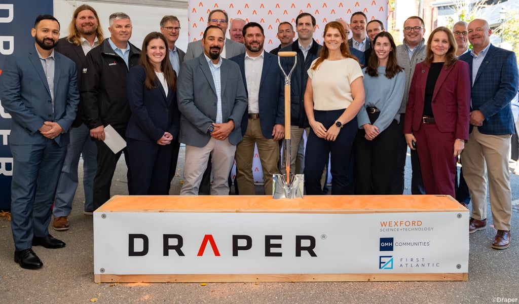 A group of individuals stands smiling in front of a backdrop displaying logos, including Draper, Wexford Science & Technology, GMH Communities, and SmartLabs, at a groundbreaking event with a silver shovel placed prominently.