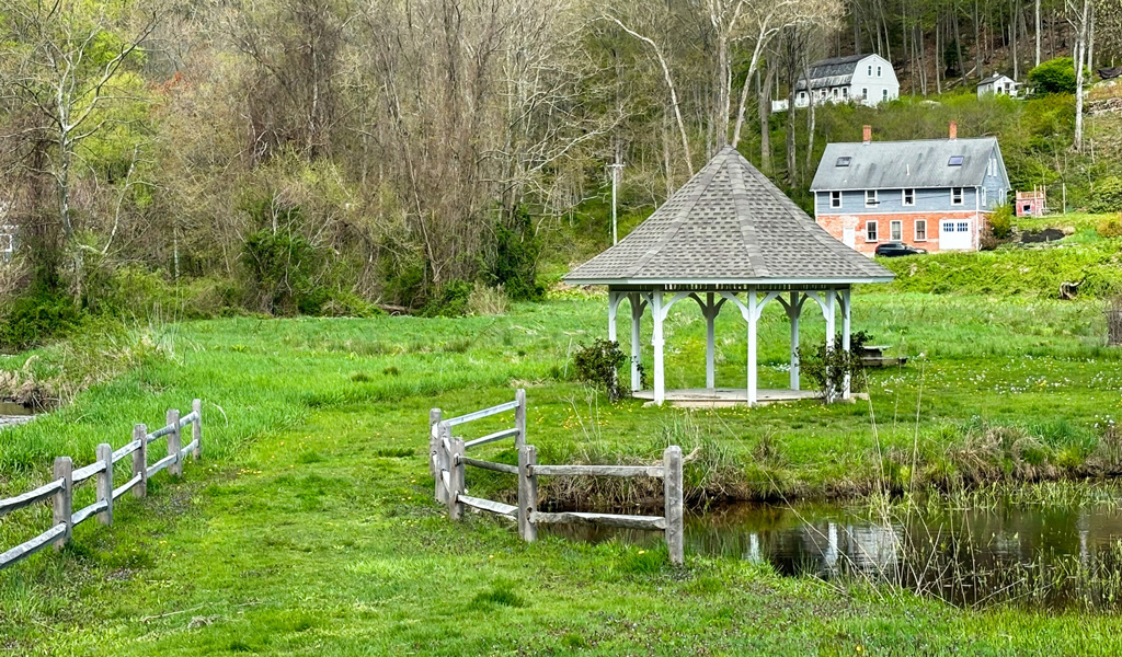 A scenic rural landscape featuring a small gazebo by a pond, with a fenced grassy field in the foreground.