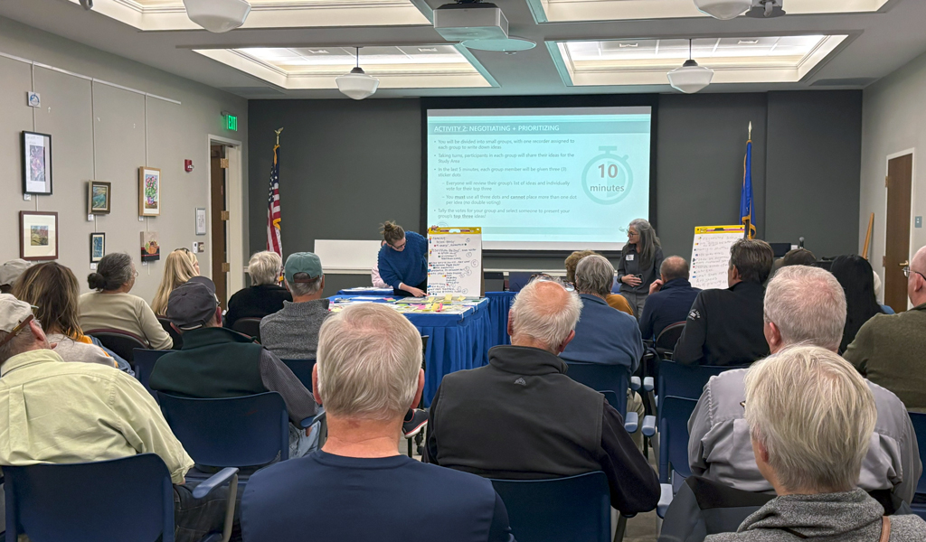 Attendees at a community meeting listen to a presentation; in the room flags and presentation materials are visible.