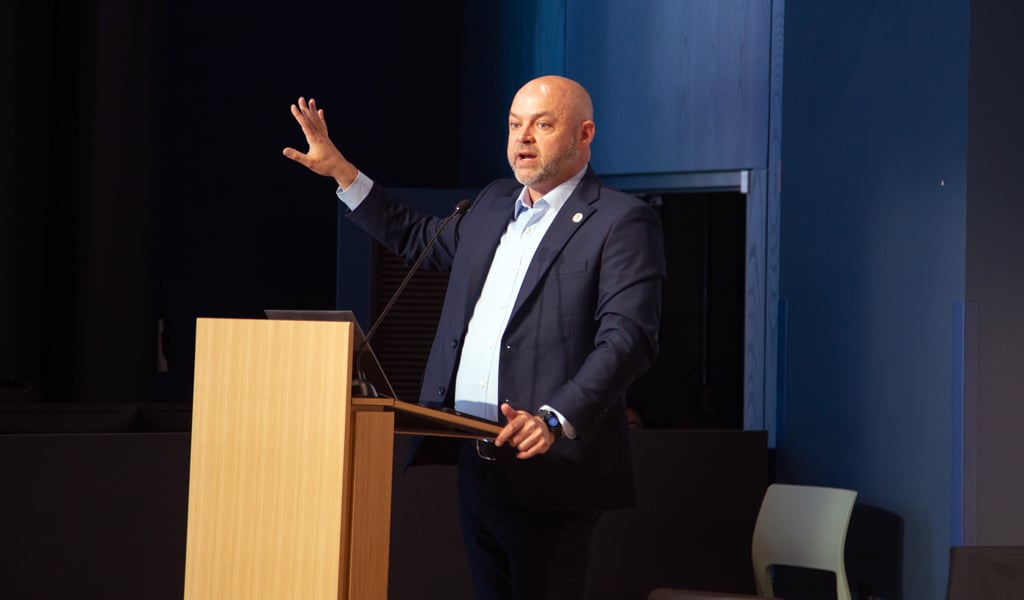 A person is speaking at a podium in a dimly lit room, raising one hand while addressing an audience.  