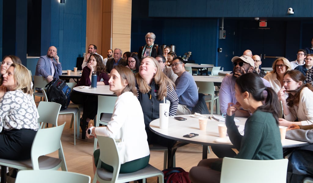 A group of people sitting at round tables in a modern conference room, attentively listening to a presentation.  