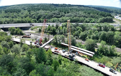Aerial view of a construction site with cranes and trucks, near a highway surrounded by green forests.