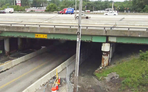 Overpass under construction with traffic cones and barriers visible, vehicles traveling on and below the overpass.