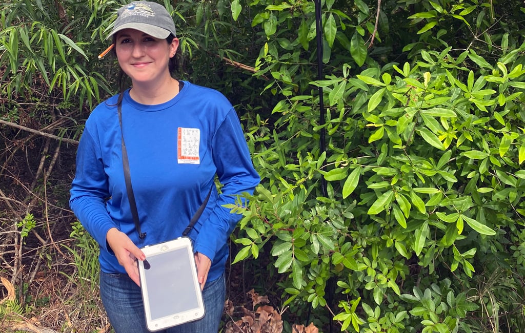 A woman in a long sleeve blue shirt, jeans, and ball cap holds an iPad and stands beside a pole used to collect acoustic data from bats