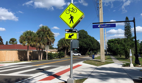 A yellow pedestrian crosswalk sign with a rectangular rapid flashing beacon and red stop sign are shown on a sunny section of the Legacy Trail 