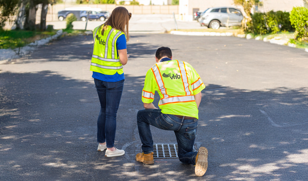 Two people wearing safety vests looking at a storm drain.