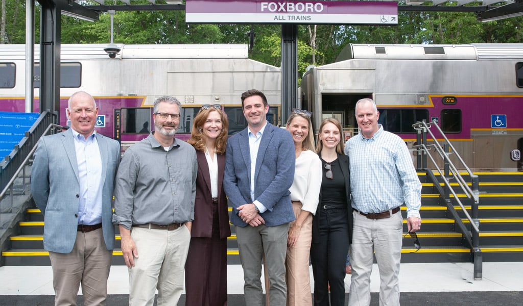 A group of smiling people in front of the Foxboro Station platform and a stationary train.