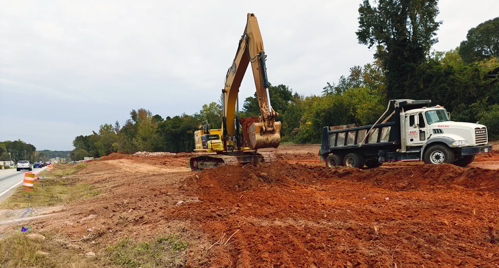 A construction excavator and dump truck beside a busy highway.