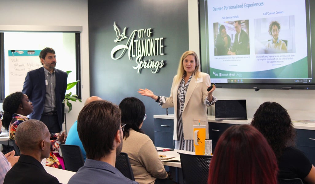 Two presenters at the front of the room in front of smart screen discussing a PowerPoint.