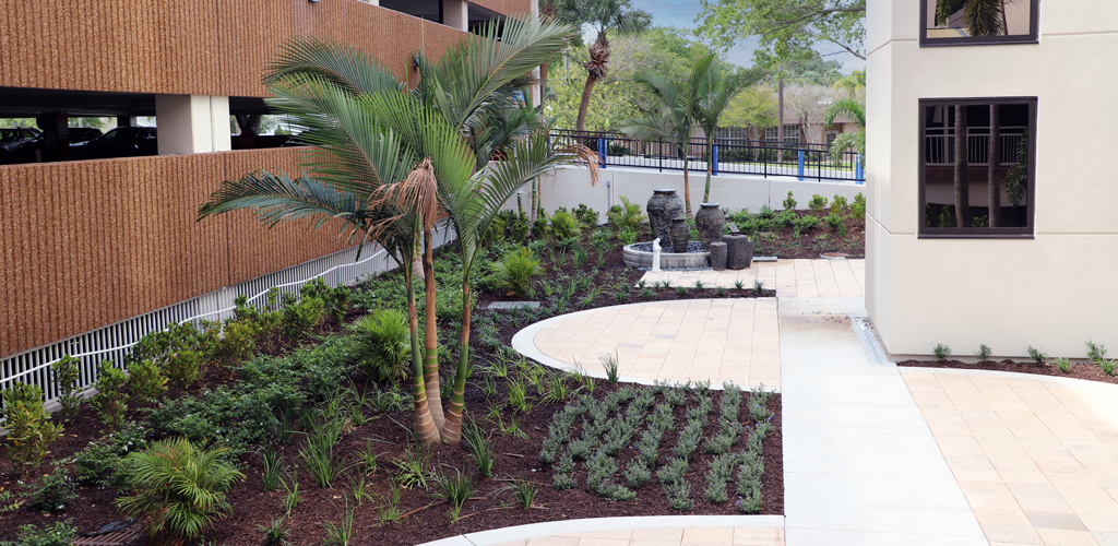 Lush tropical landscaping and a fountain in between a modern hospital and partking garage. 