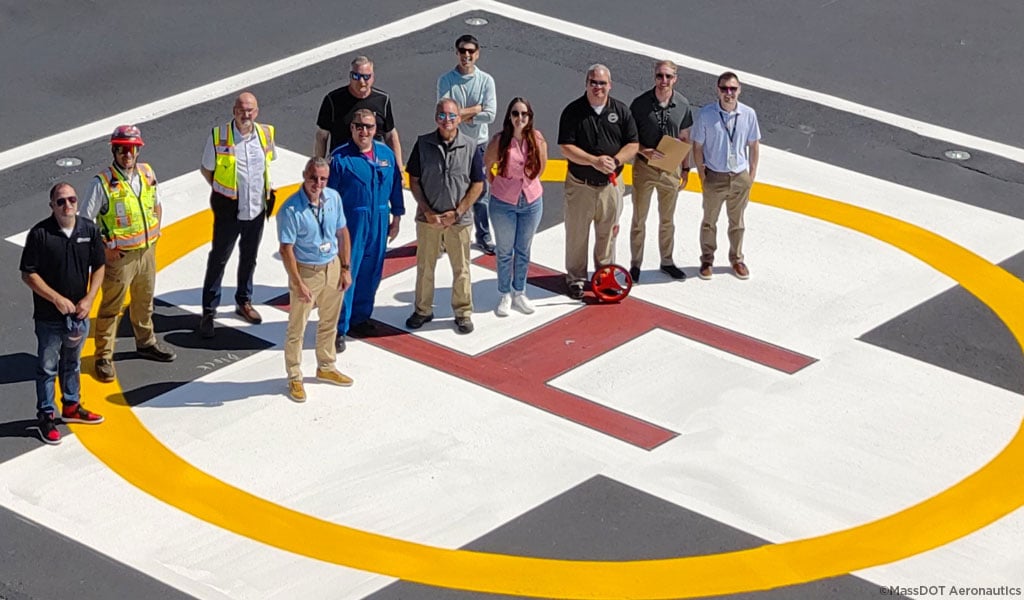 Aerial image of the project team smiling on a helipad with the hospital behind them.