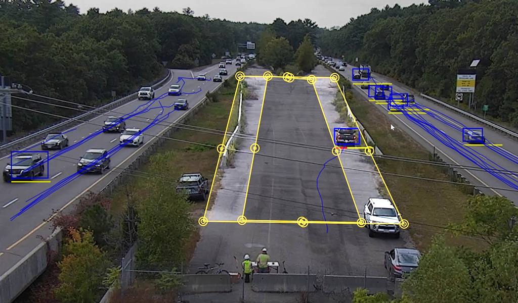 This image shows a road with multiple lanes of traffic, featuring several cars and a truck. Yellow and blue lines are overlaid, possibly indicating traffic monitoring or analysis. 