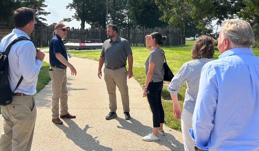 A group of people stand on a paved pathway at Jamestown Island, discussing site conditions during an outdoor project visit.