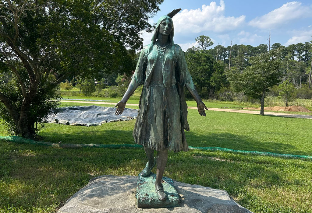 A bronze statue of Pocahontas stands on a stone base beside a paved pathway at Jamestown Island. 