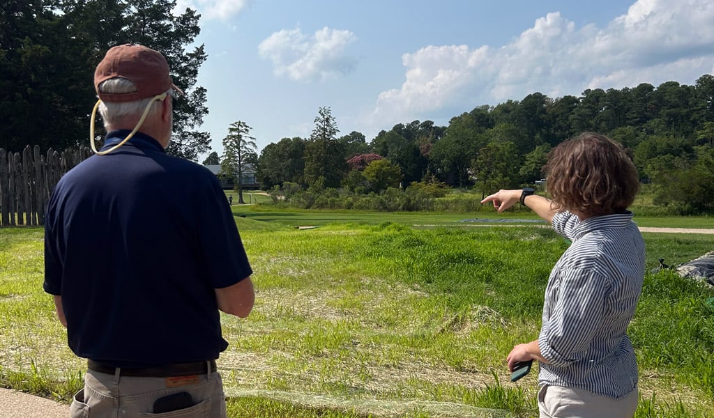 Two people stand beside a paved walkway overlooking an open grassy area at Jamestown Island. One person gestures toward the landscape while discussing conditions across the site.