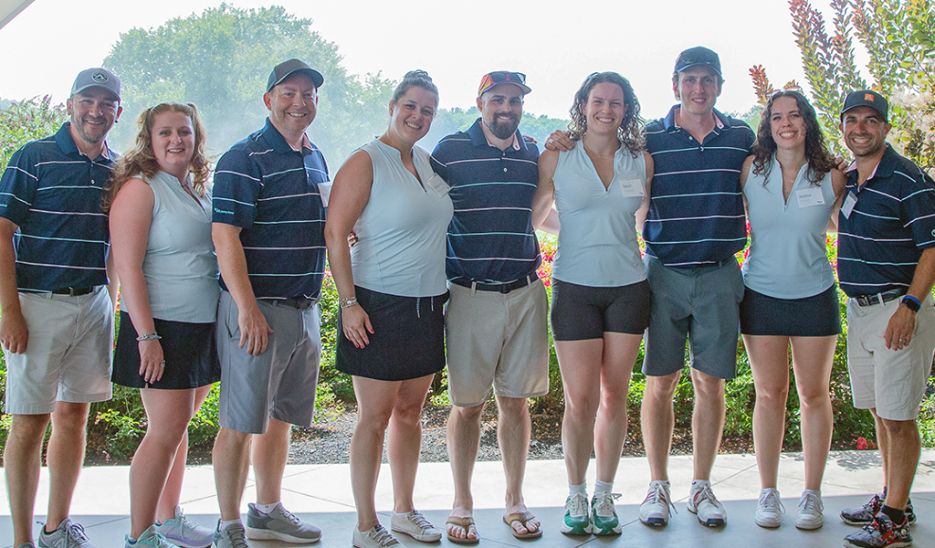 A group of eight individuals standing together inside a patio area, all dressed in navy blue and white golf attire, with a background of colorful plants and trees bathed in soft daylight.
