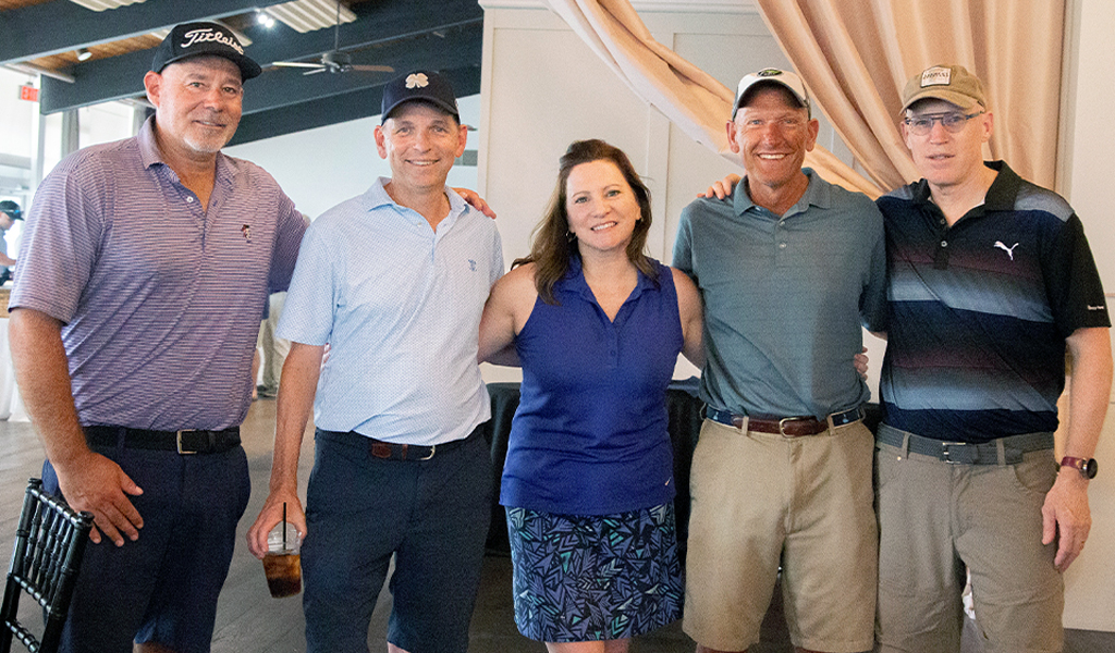 A group of five individuals standing together in an indoor setting. They are casually dressed in golf attire, with several wearing caps.