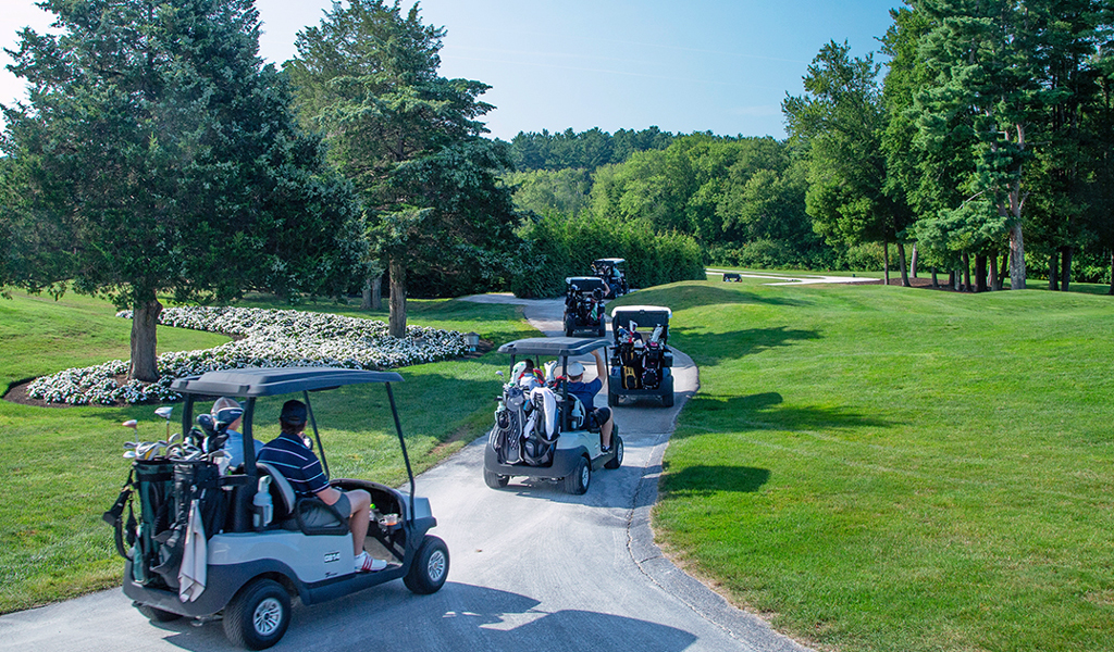 A line of golf carts carrying golfers and their equipment winds along a paved path through a golf course.