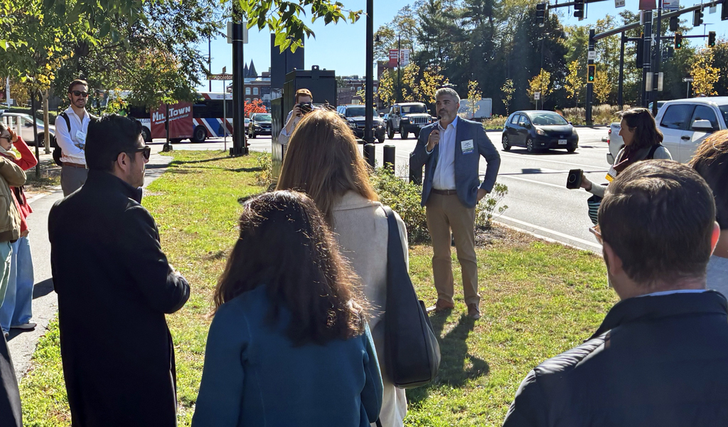 Group of people listening to a speaker at an outdoor event near a busy intersection.