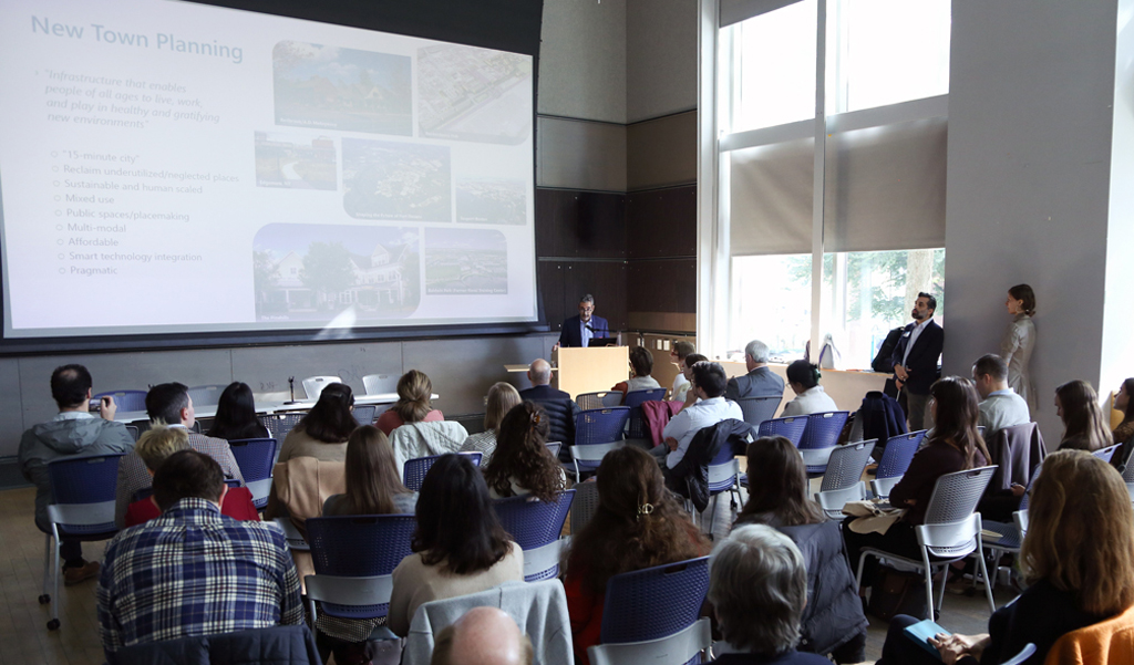 Audience attending a presentation on "New Town Planning" in a lecture hall, with a speaker at a podium displaying slides.