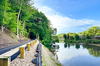 Paved road running alongside a calm body of water, bordered by trees, guardrails, and a gravel shoreline.