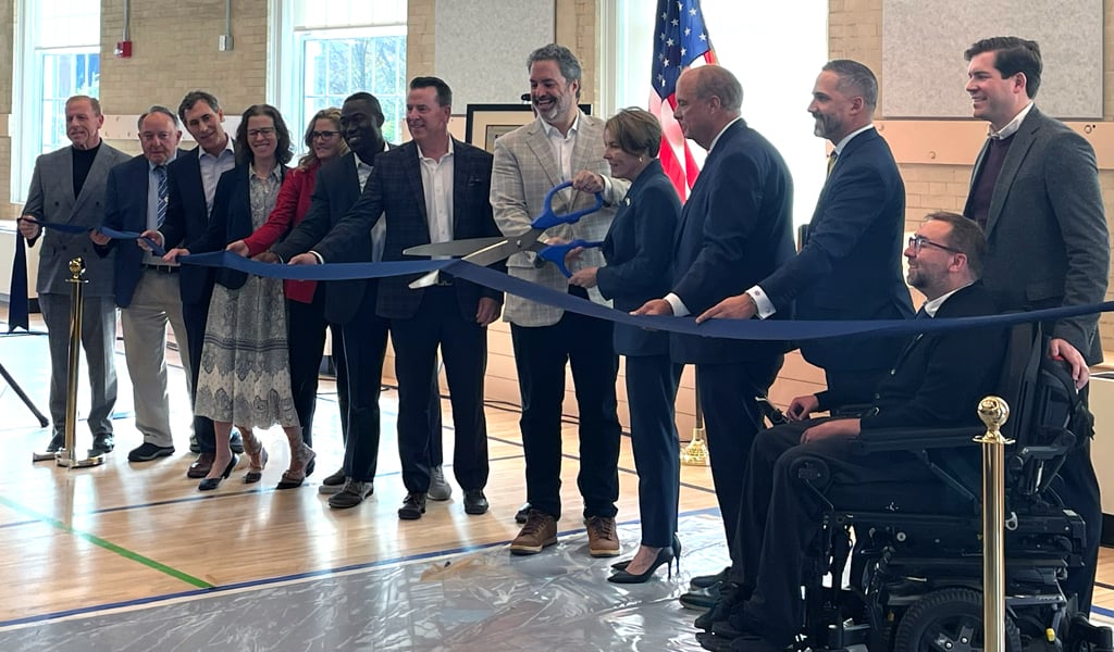 A group of people stands on an indoor stage gathered for a ribbon-cutting ceremony. The person in the middle cuts the ribbon with oversized scissors. 