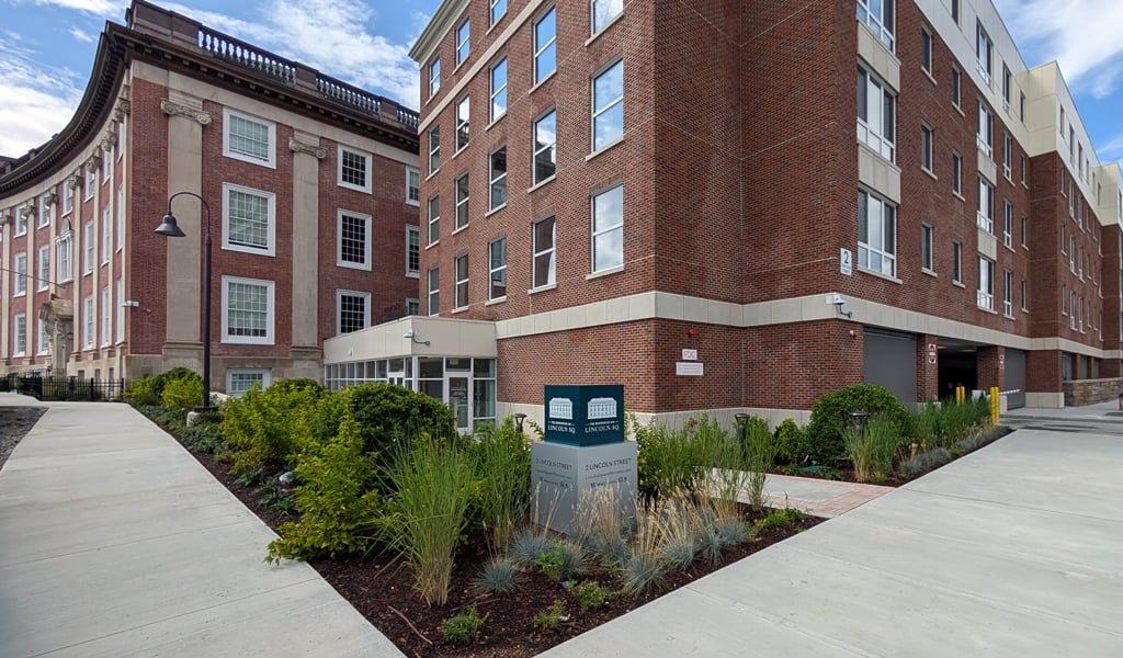 A large brick building with multiple stories is situated on a corner. The building is adjacent to a smaller brick structure with a rounded facade.  