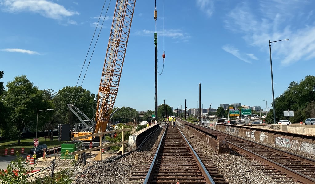 Construction equipment operates along a river beside a concrete bridge, with workers and materials visible under cloudy sky.